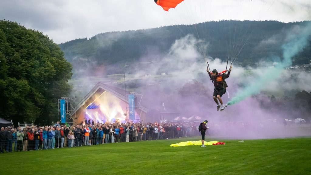 En faldskærmsudspringer lander med røg i baggrunden ved en udendørs event, mens en stor menneskemængde ser til.